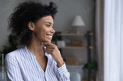 Smiling young African American woman look in window distance dreaming or visualizing perspectives. Happy mixed race female dream or make plans decision, imagine opportunities. Vision concept.