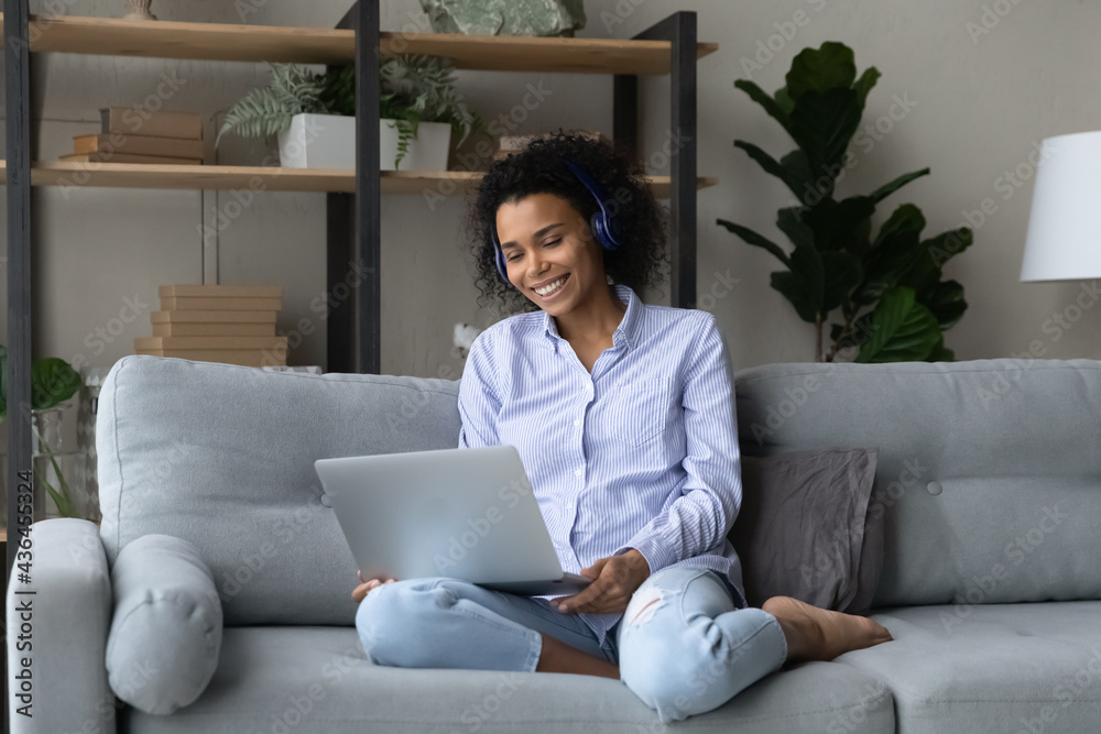 Smiling young African American woman in earphones relax on sofa at home use modern laptop gadget. Happy millennial mixed race female in headphones talk speak on webcam video call on computer. Smiling young African American woman in earphones relax on sofa at home use modern laptop gadget. Happy millennial mixed race female in headphones talk speak on webcam video call on computer.