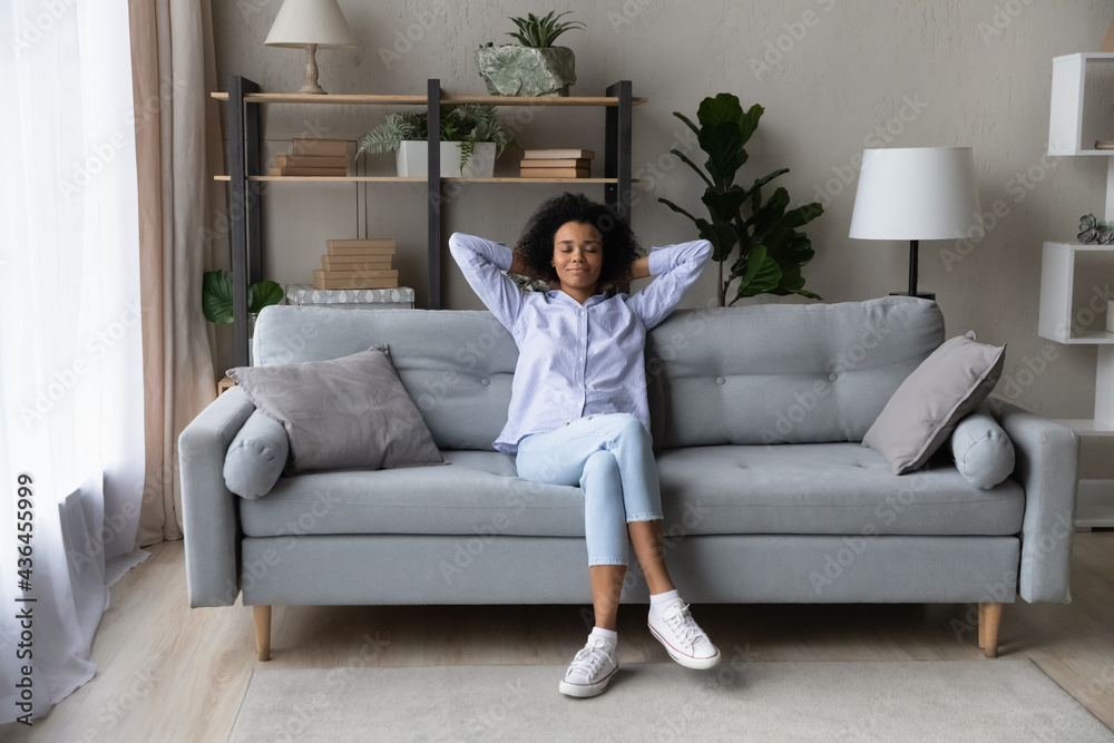Happy calm young African American woman sit relax on sofa in living room take nap or sleep. Smiling millennial mixed race female renter rest on couch at home breathe fresh air. Rental concept. Happy calm young African American woman sit relax on sofa in living room take nap or sleep. Smiling millennial mixed race female renter rest on couch at home breathe fresh air. Rental concept.