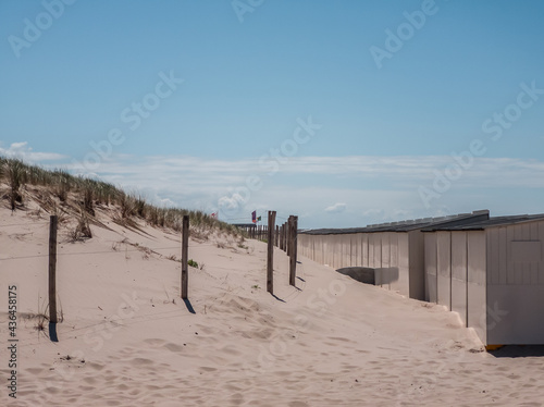 A fence with wooden poles and beach houses in front of a dune with beach grass and blue sky as background
