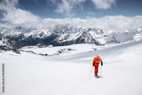 Young male freerider guide in a yellow ski suit with a backpack on his shoulders climbing the mountain on skis on a sunny day. © yanik88