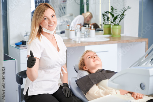Cheerful female stomatologist showing approval gesture and smiling while woman lying in dental chair. Joyful dentist in sterile gloves doing thumbs up while sitting beside patient in dental office.