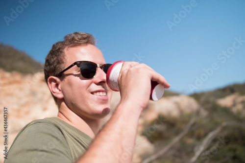 Happy young man wearing sunglasses and drinking coffe on a sunny day in the mountain. © Max Roquer