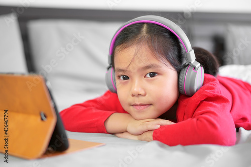 Asian child or kid girl learn from home or people study online on computer tablet and wearing headphone to e-learning communication on pad to smiling and sleep lie on stomach on bed by back to school