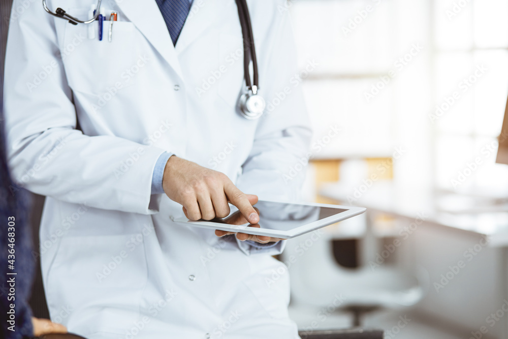 Unknown red-bearded doctor and patient-woman discussing current health examination while sitting and using tablet computer in sunny clinic, close-up. Medicine concept Unknown red-bearded doctor and patient-woman discussing current health examination while sitting and using tablet computer in sunny clinic, close-up. Medicine concept
