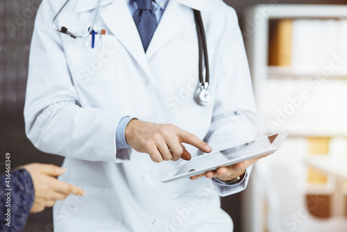 Unknown red-bearded doctor and patient-woman discussing current health examination while sitting and using tablet computer in sunny clinic, close-up. Medicine concept
