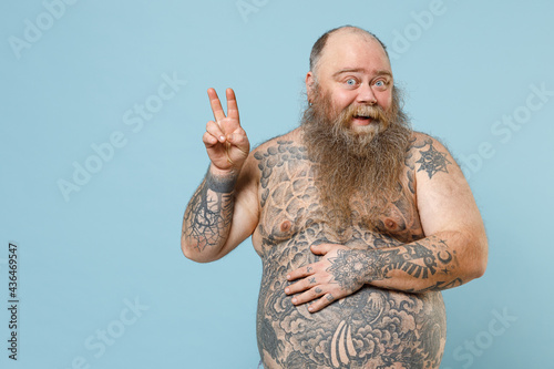 Young fat pudge obese chubby overweight blue-eyed bearded man has big belly with naked tattooed torso show v-sign victory gesture isolated on pastel blue background studio portrait studio portrait photo