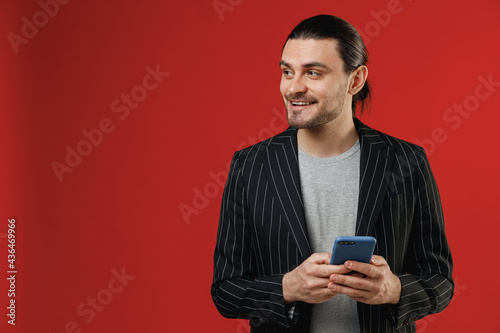 Young surprised businessman latin man with ponytail in black striped jacket grey shirt point index finger on mobile cell phone browsing surfing internet look aside isolated on red color background