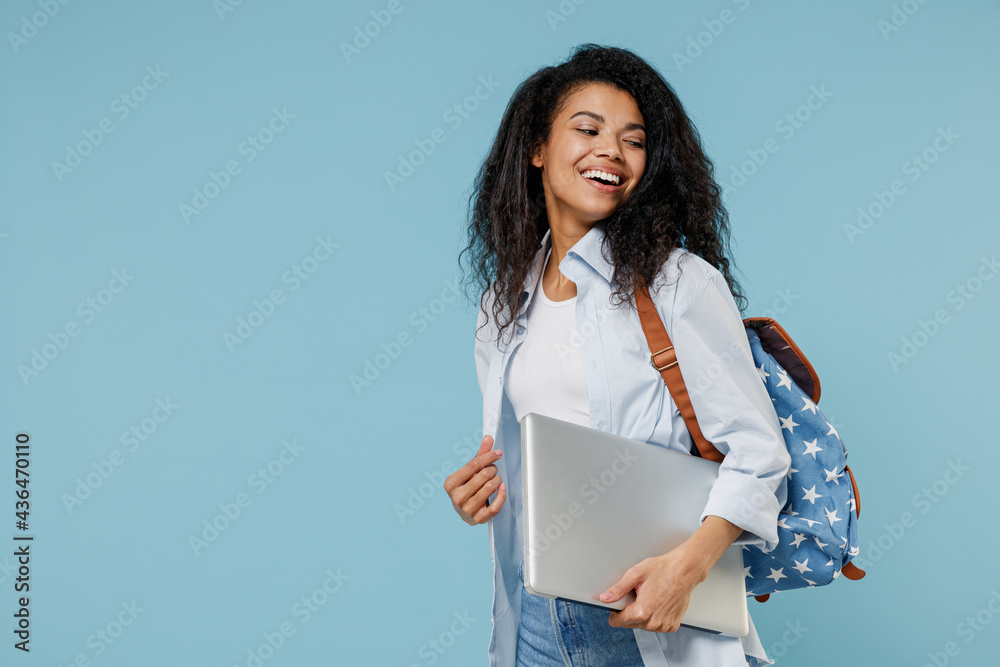 Obraz premium Young smiling african american girl teen student in denim clothes backpack hold closed laptop pc computer look aside isolated on blue background Education in high school university college concept.