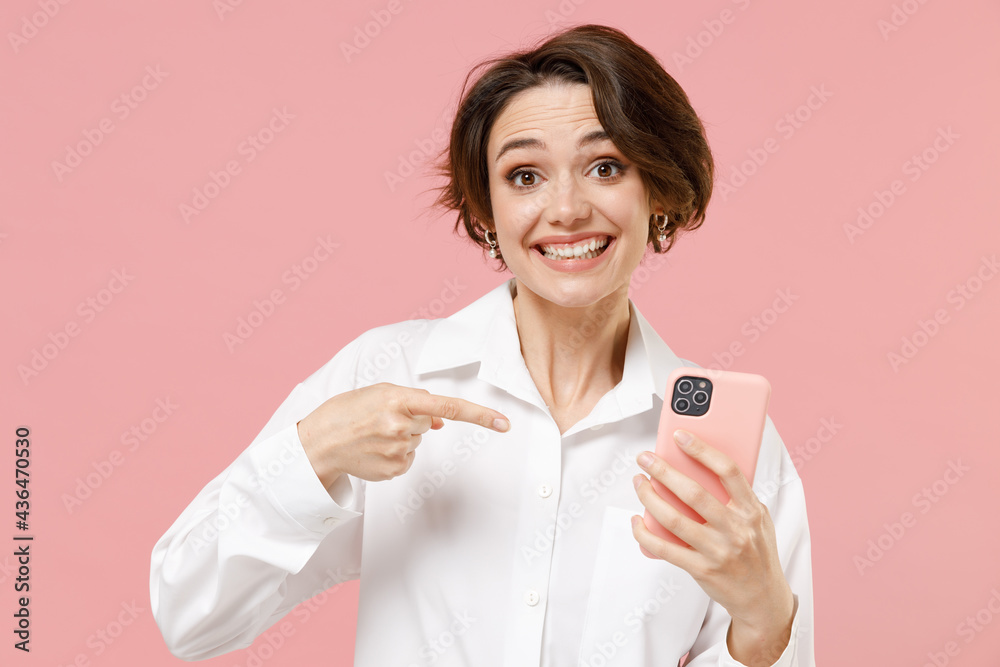 Amazed fun young employee business woman corporate lawyer in classic formal white shirt work in office hold point index finger mobile cell phone chat online isolated on pastel pink background studio. Amazed fun young employee business woman corporate lawyer in classic formal white shirt work in office hold point index finger mobile cell phone chat online isolated on pastel pink background studio.