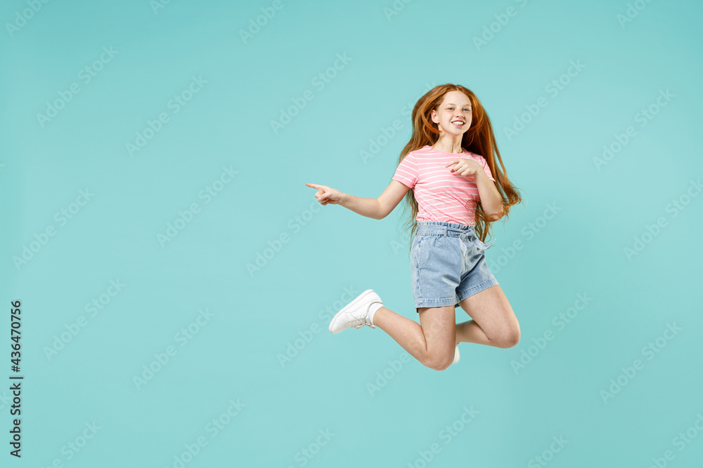 Full length little redhead kid girl 12-13 year old in pink striped t-shirt jump point index finger aside on workspace mock up isolated on pastel blue background Children lifestyle childhood concept.