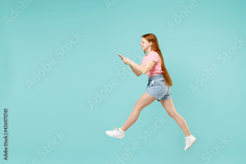 Fototapeta Naklejka Na Ścianę i Meble -  Full length little side view funny redhead kid girl 12-13 years old in pink t-shirt hold mobile cell phone jump high isolated on pastel blue background studio. Children lifestyle childhood concept