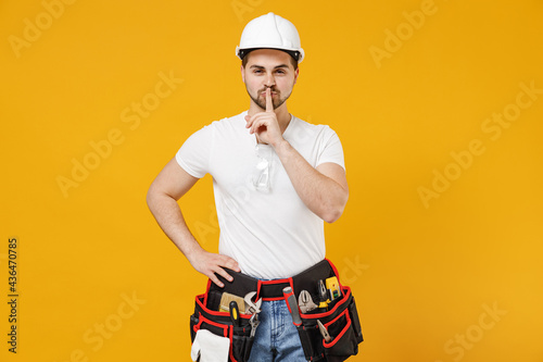 Young employee handyman man in protective helmet hardhat say hush quiet finger on lips shh gesture isolated on yellow background Instruments accessories renovation apartment room Repair home concept.