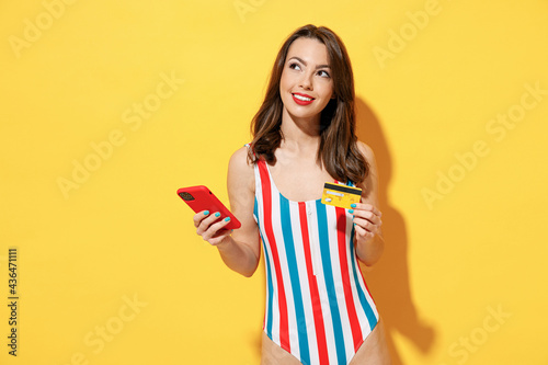 Happy young sexy woman slim body wear red blue swimsuit using mobile cell phone hold credit bank card isolated on vivid yellow color wall background studio. Summer hotel pool sea rest sun tan concept. photo