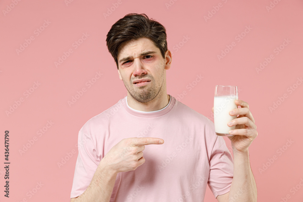 Sick sad unhealthy ill allergic man has red watery eyes, runny stuffy sore nose suffer from intolerance lactose allergy trigger symptom hold milk glass isolated on pastel pink color background studio