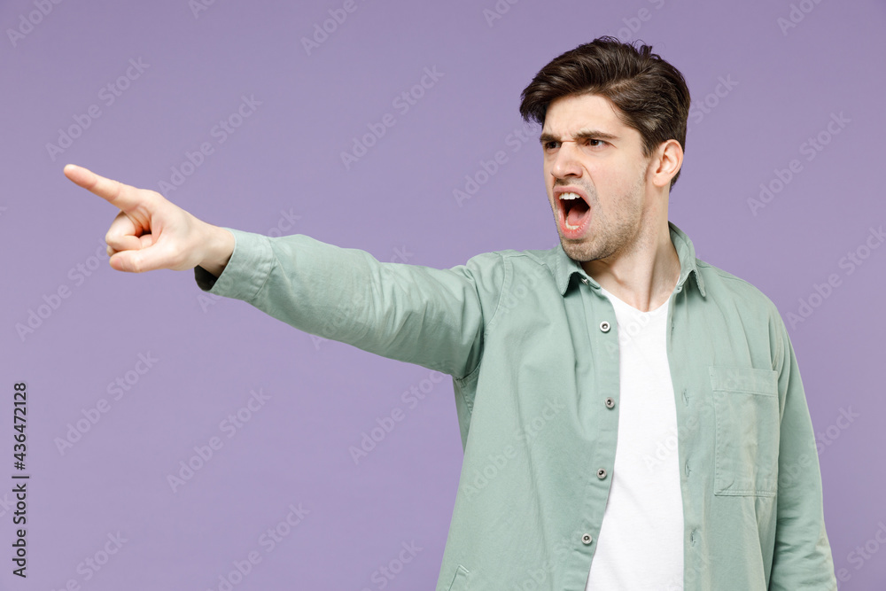 Young angry indignant sad displeased caucasian man 20s in casual mint shirt white t-shirt point index finger aside scream shout isolated on purple background studio portrait. People lifestyle concept Young angry indignant sad displeased caucasian man 20s in casual mint shirt white t-shirt point index finger aside scream shout isolated on purple background studio portrait. People lifestyle concept