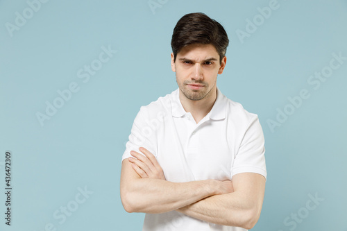 Young sad disappointed unshaven caucasian man 20s wearing white casual basic t-shirt hold hands crossed folded look camera isolated on pastel blue background studio portrait. People lifestyle concept.