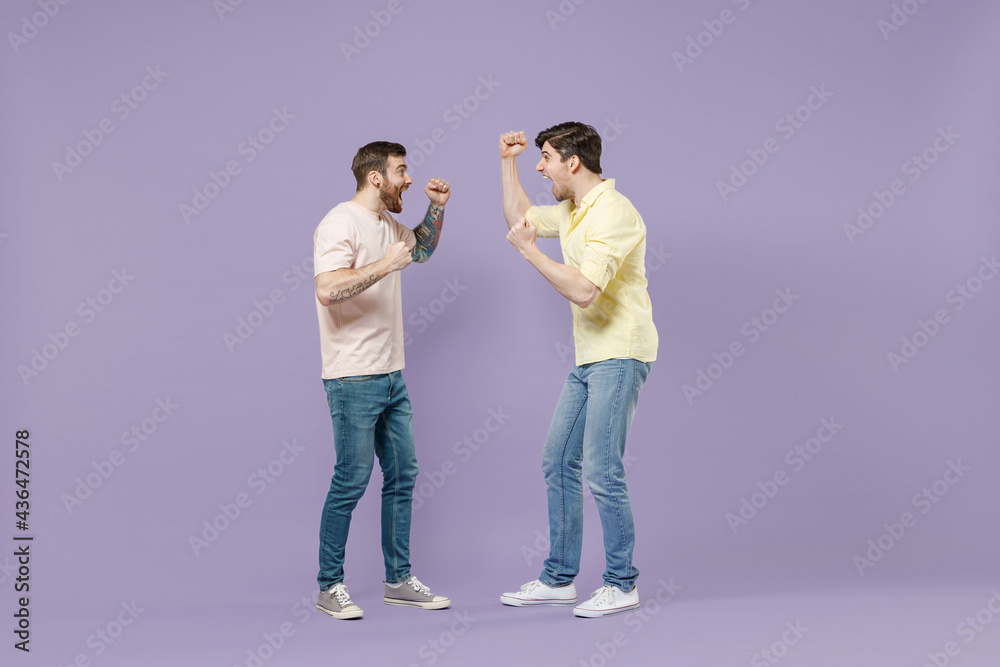 Full length overjoye two young happy men friends together walk in casual t-shirt walk do winner gesture clench fist look to each other isolated on purple background People friendship lifestyle concept