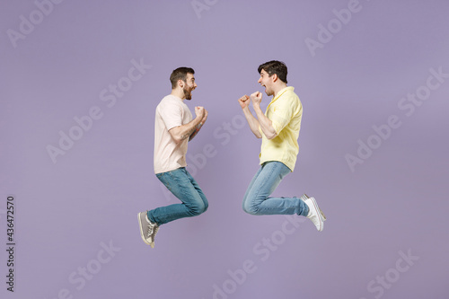 Side view full length two young happy men friends together in casual t-shirt looking to each other do winner gesture clench fist jump high isolated on purple background studio People lifestyle concept