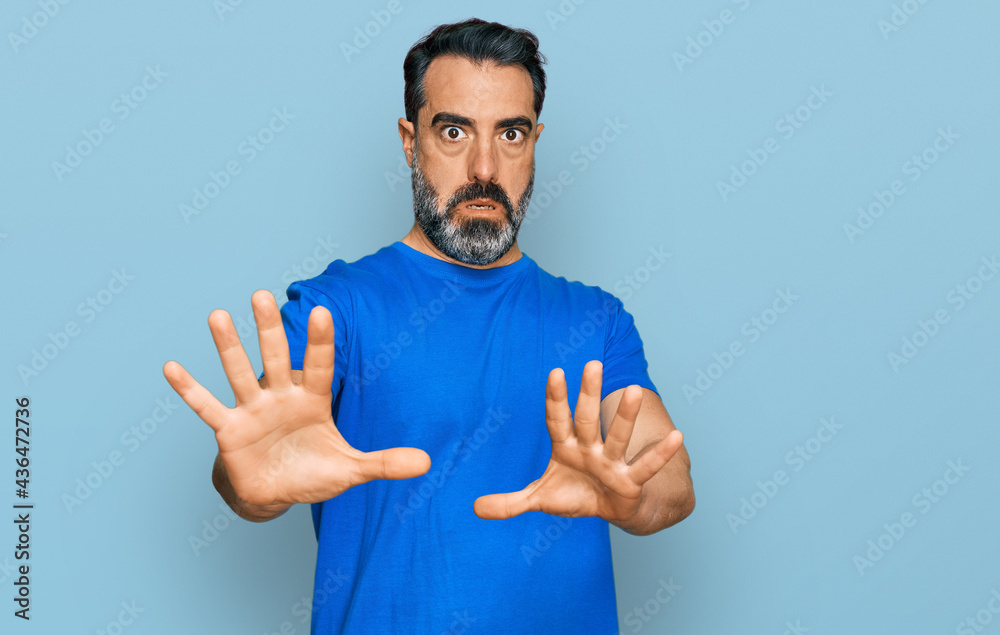 Middle aged man with beard wearing casual blue t shirt afraid and terrified with fear expression stop gesture with hands, shouting in shock. panic concept. Middle aged man with beard wearing casual blue t shirt afraid and terrified with fear expression stop gesture with hands, shouting in shock. panic concept.
