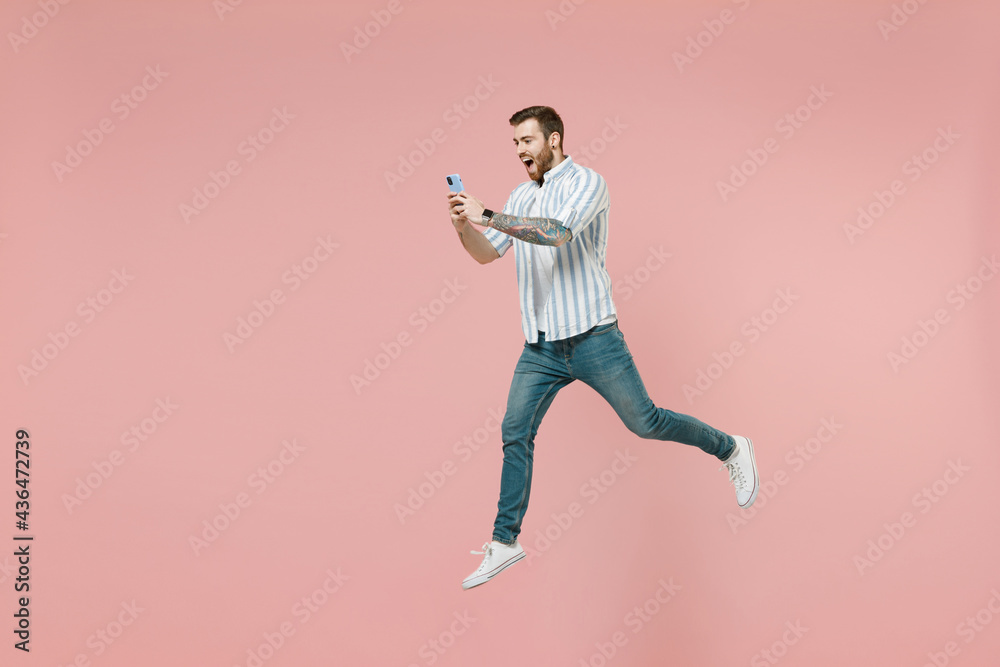 Full length young overjoyed unshaven man 20s wear blue striped shirt white t-shirt jump high hold mobile cell phone chat online isolated on pastel pink background studio portrait Tattoo translate fun