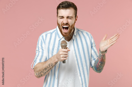 Young expressive cool unshaven man 20s wearing blue striped shirt sing song in microphone at karaoke club spread hands isolated on pastel pink color background studio portrait. Tattoo translate fun.