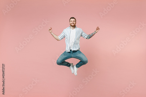 Full length young unshaven man 20s wearing blue striped shirt white t-shirt jump high hold spreading hands in yoga om aum gesture relax meditate try to calm down isolated on pastel pink background