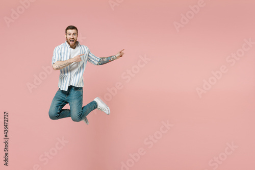 Full length young amazed unshaven man wear blue striped shirt white t-shirt jump high point index finger aside on copy space workspace area isolated on pastel pink background. Tattoo translate fun