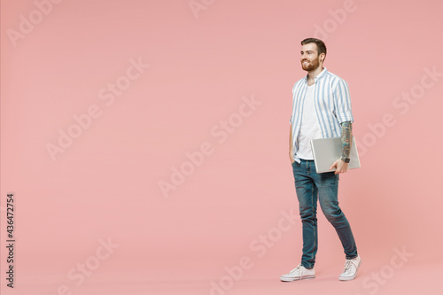 Full length young smiling successful unshaven man in blue striped shirt holding closed laptop pc computer walking going look aside isolated on pastel pink background studio People lifestyle concept.