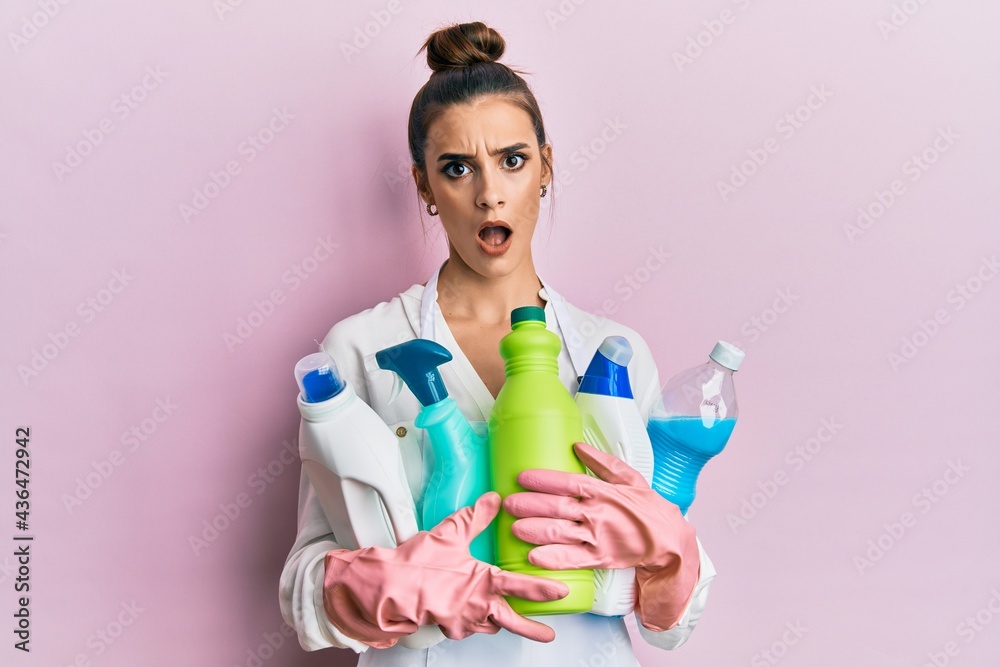 Beautiful brunette young woman wearing cleaner apron holding cleaning products in shock face, looking skeptical and sarcastic, surprised with open mouth