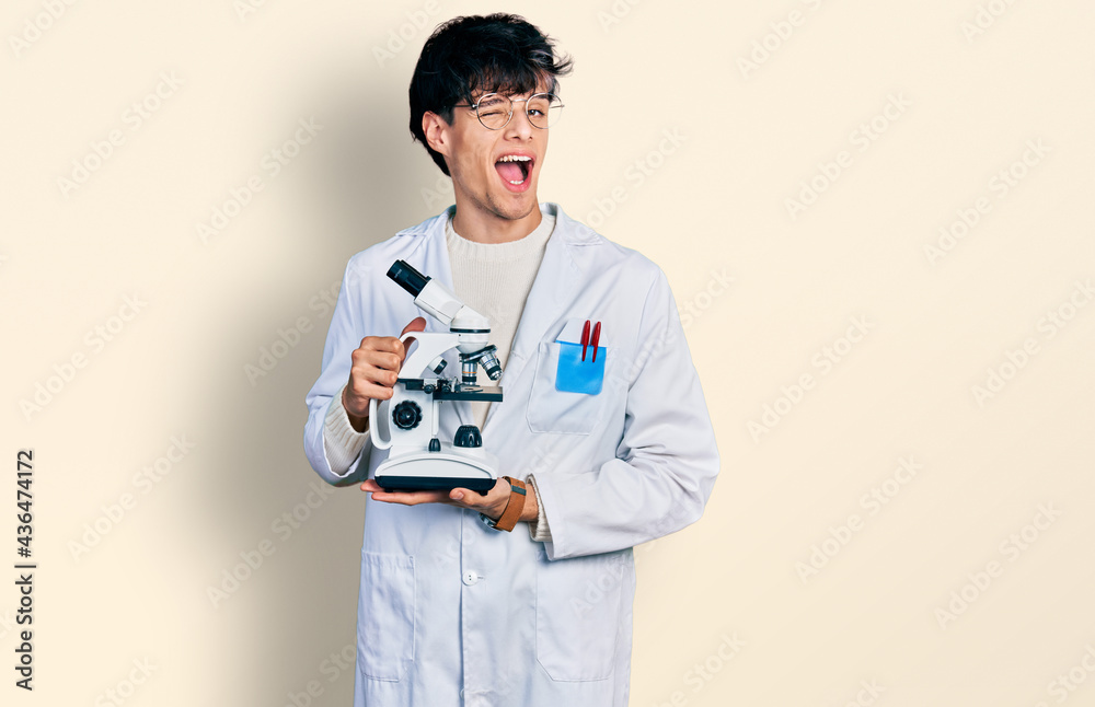 Handsome hipster young man wearing lab coat holding microscope winking looking at the camera with sexy expression, cheerful and happy face.