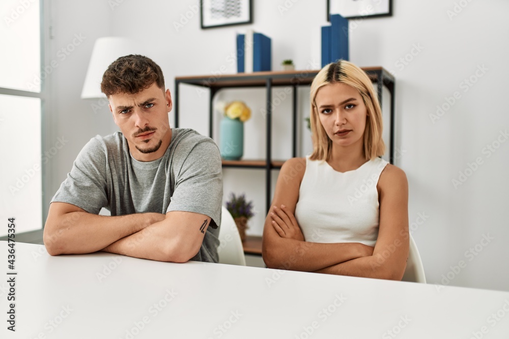 Young beautiful couple wearing casual clothes sitting on the table at home skeptic and nervous, disapproving expression on face with crossed arms. negative person. Young beautiful couple wearing casual clothes sitting on the table at home skeptic and nervous, disapproving expression on face with crossed arms. negative person.