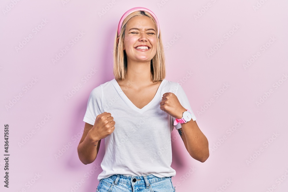 Beautiful blonde woman wearing casual white t shirt very happy and excited doing winner gesture with arms raised, smiling and screaming for success. celebration concept. Beautiful blonde woman wearing casual white t shirt very happy and excited doing winner gesture with arms raised, smiling and screaming for success. celebration concept.