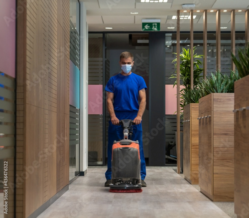Cleaner with the mask on his face cleans the hard floor in the office with walk behind scrubber dryer machine.Cleaning and maintenance concept
