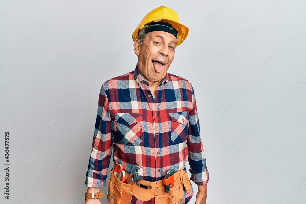 Senior hispanic man wearing handyman uniform sticking tongue out happy with funny expression. emotion concept.