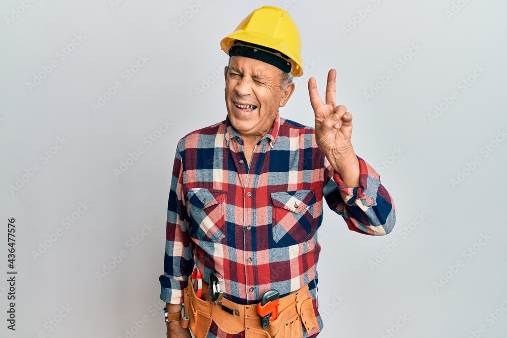 Senior hispanic man wearing handyman uniform smiling with happy face winking at the camera doing victory sign. number two. Senior hispanic man wearing handyman uniform smiling with happy face winking at the camera doing victory sign. number two.