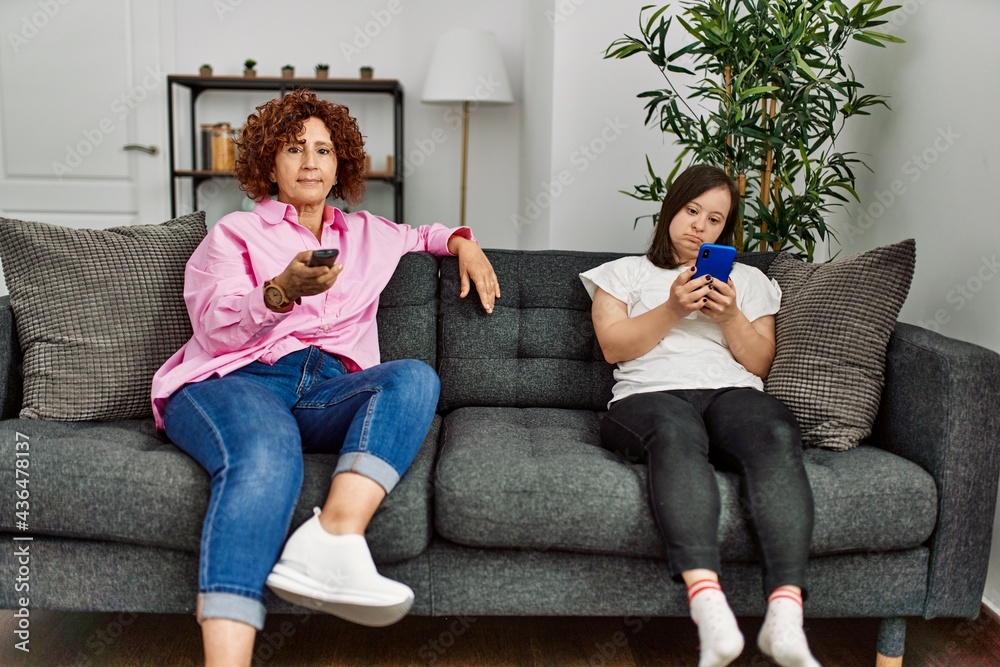 Obraz premium Mature mother and down syndrome daughter at home watching television and using smartphone