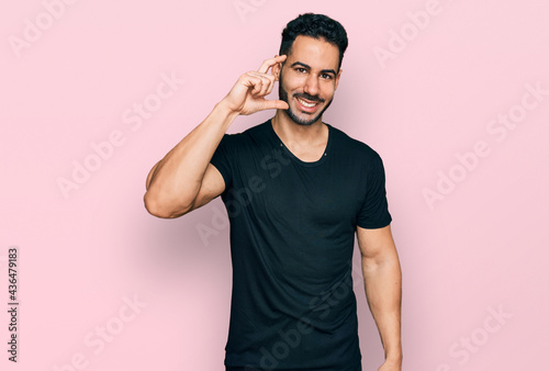 Hispanic man with beard wearing casual black t shirt smiling and confident gesturing with hand doing small size sign with fingers looking and the camera. measure concept.