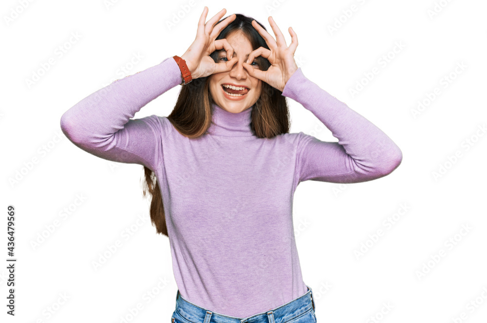 Young beautiful teen girl wearing turtleneck sweater doing ok gesture like binoculars sticking tongue out, eyes looking through fingers. crazy expression. Young beautiful teen girl wearing turtleneck sweater doing ok gesture like binoculars sticking tongue out, eyes looking through fingers. crazy expression.