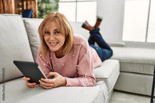 Middle age blonde woman using touchpad lying on the sofa at home.