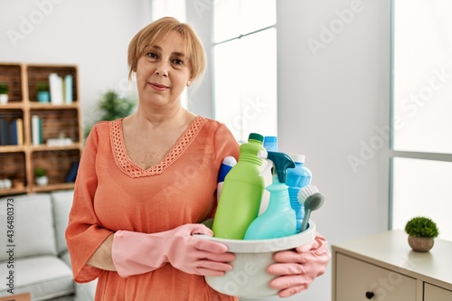 Middle age blonde woman holding cleaning products cleaning at home thinking attitude and sober expression looking self confident