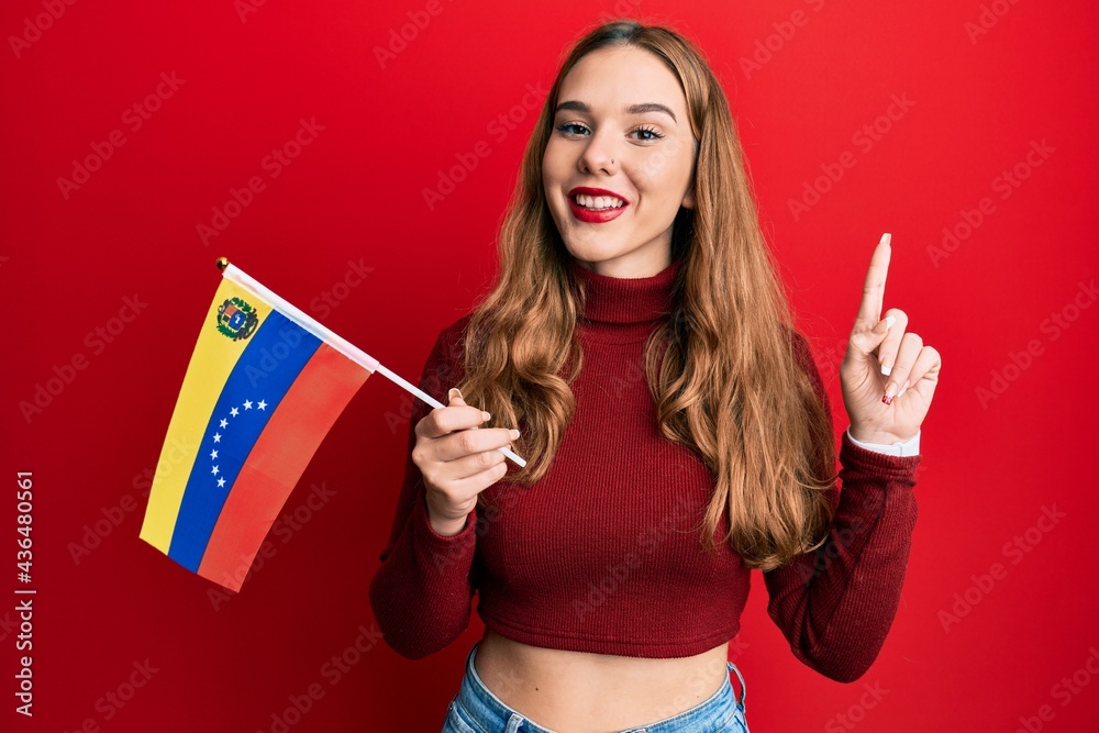Young blonde woman holding venezuelan flag smiling with an idea or question pointing finger with happy face, number one