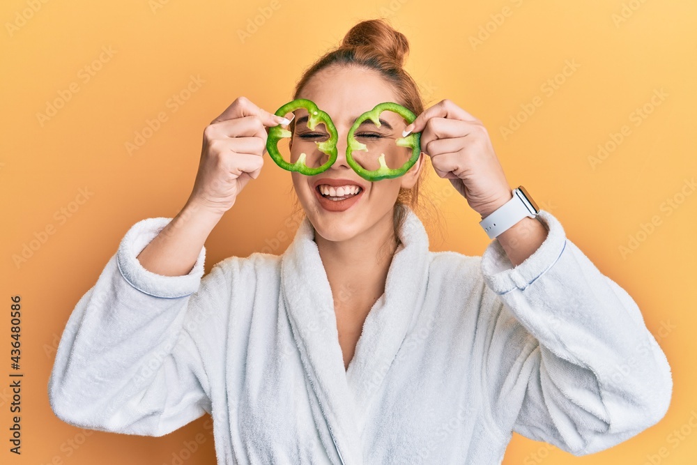 Young blonde woman wearing bathrobe holding green pepper over eyes smiling and laughing hard out loud because funny crazy joke. Young blonde woman wearing bathrobe holding green pepper over eyes smiling and laughing hard out loud because funny crazy joke.