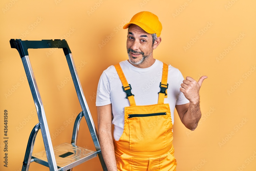 Handsome middle age man with grey hair holding ladder smiling with happy face looking and pointing to the side with thumb up.