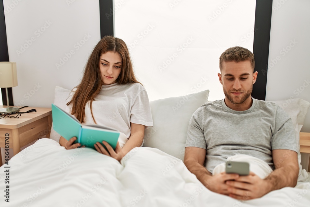 Young caucasian couple reading book and using smartphone at bed. Young caucasian couple reading book and using smartphone at bed.