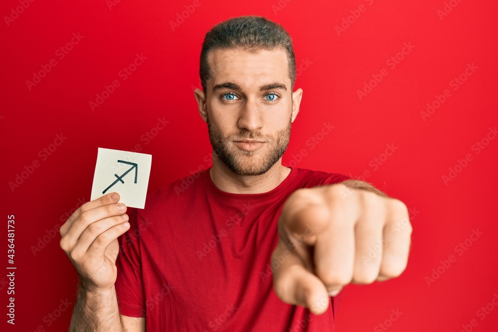 Young caucasian man holding paper with sagittarius zodiac sign pointing with finger to the camera and to you, confident gesture looking serious Young caucasian man holding paper with sagittarius zodiac sign pointing with finger to the camera and to you, confident gesture looking serious