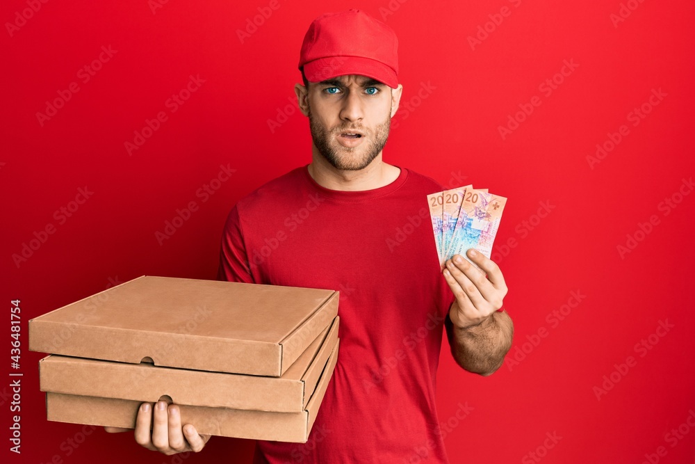 Young caucasian man holding delivery box and swiss franc banknotes in shock face, looking skeptical and sarcastic, surprised with open mouth Young caucasian man holding delivery box and swiss franc banknotes in shock face, looking skeptical and sarcastic, surprised with open mouth