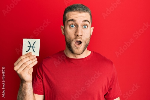 Young caucasian man holding paper with pisces zodiac sign scared and amazed with open mouth for surprise, disbelief face