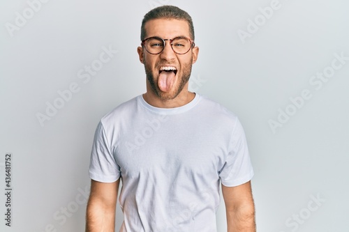 Young caucasian man wearing casual clothes and glasses sticking tongue out happy with funny expression. emotion concept.