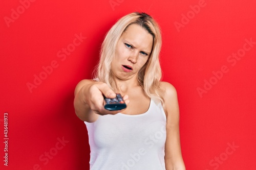 Beautiful caucasian blonde woman holding television remote control in shock face, looking skeptical and sarcastic, surprised with open mouth